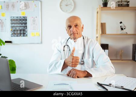 mature doctor with stethoscope works on a laptop looking at camera approving doing positive gesture with hand, thumb-up. Senior therapist making video Stock Photo
