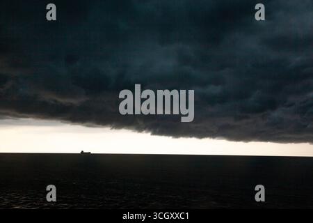 A stormy sky in the sea with thick dark clouds and a bright gap between the sky and the sea in which a cargo ship can be seen. Stock Photo