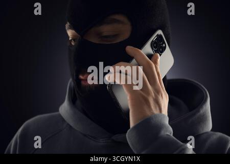 Criminal in mask talking on phone against dark background Stock Photo