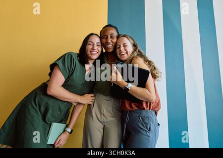 Diverse young businesswomen hugging and smiling while standing against colorful wall in office space Stock Photo