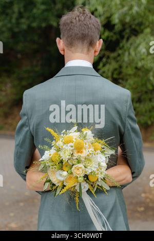 Man in a suit conceals a bouquet of yellow and white flowers behind his back, standing outdoors. The bouquet features roses and daisies, adding a Stock Photo