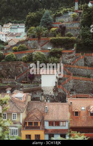 Vertical shot of a terraced hillside gardens in Cudillero, Spain, with stone walls, stairways, and lush greenery rising above colorful village houses Stock Photo