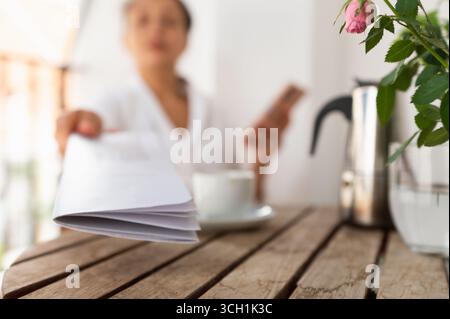 A close-up view of a hand extending a document across a wooden table with coffee, a metallic pitcher, and a flower vase in focus. The background shows Stock Photo
