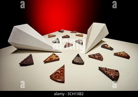 An artistic layout of broken chocolate bars spread across a white background, arranged with geometric prism structures, against a red illuminated back Stock Photo