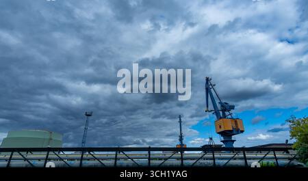 Cranes and storage tanks at an industrial power facility under a moody cloudy sky, captured in a wide-angle landscape view. Stock Photo