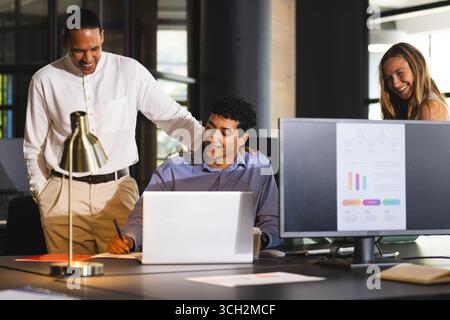Diverse colleagues collaborating at office, smiling and discussing project on laptop Stock Photo