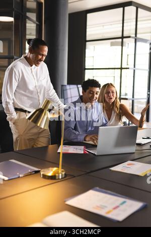 Collaborating in modern office, diverse colleagues sharing ideas and laughing over laptop Stock Photo