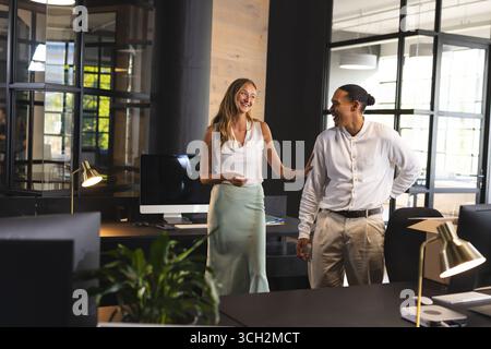 Diverse colleagues laughing and chatting in modern office with computers and plants Stock Photo
