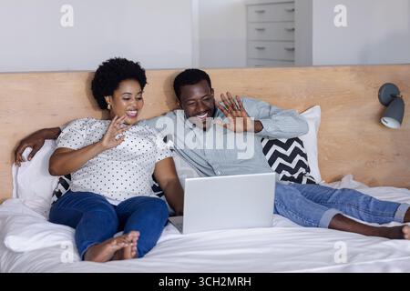 Couple relaxing on bed, video chatting with friends on laptop, smiling happily Stock Photo