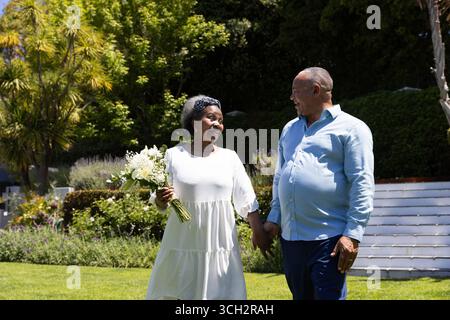 African American senior couple walking in garden, holding hands and smiling warmly Stock Photo