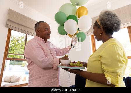 Senior couple enjoying balloons and fruit platter at home, sharing joyful moment Stock Photo