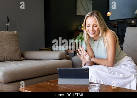 Woman in twenties leaning toward tablet on light-gray sofa in modern living room, copy space Stock Photo