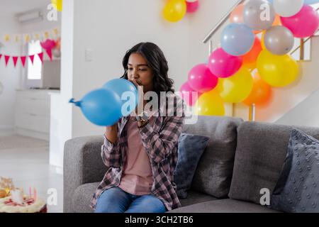 Woman inflating blue balloon on gray couch in decorated living room, with birthday cake and bunting Stock Photo