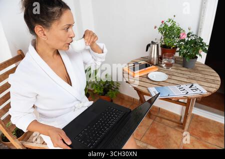A focused woman sits on a patio, holding a coffee cup while working on her laptop. Decorated with plants, the setting exudes a relaxed yet productive Stock Photo