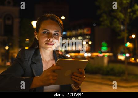 Young adult female with digital tablet and earbuds on evening city street Stock Photo