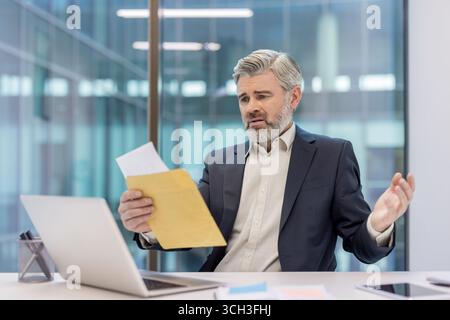 A mature businessman looks concerned while reviewing paperwork in a modern office setting. Stock Photo