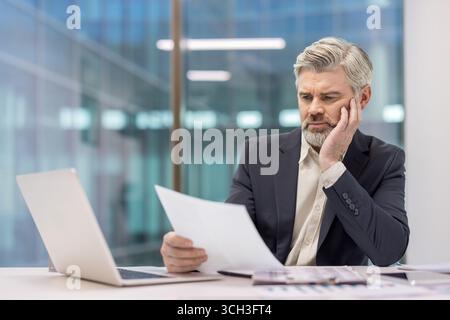 A senior professional examines paperwork in a modern office environment, conveying analysis and professionalism. Stock Photo