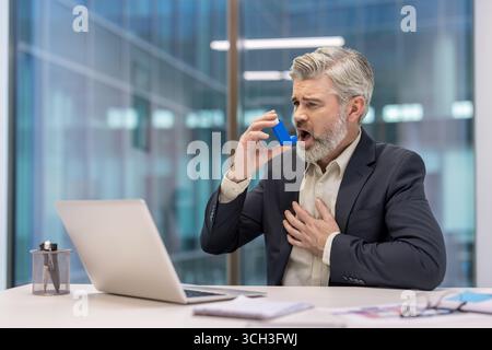 Middle-aged man in business attire using an inhaler, sitting at his desk in a modern office. The image conveys health concerns, respiratory issues, and professional settings. Stock Photo