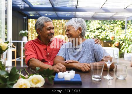 Elderly diverse couple sitting at table, smiling and enjoying time together in garden Stock Photo