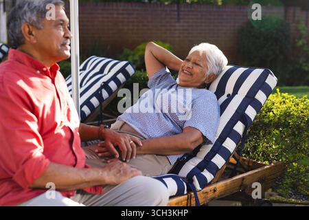 Senior diverse couple relaxing on patio chairs, enjoying conversation in sunny backyard Stock Photo