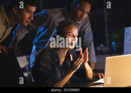 Diverse team collaborating on laptop at night, woman using headset for video call Stock Photo