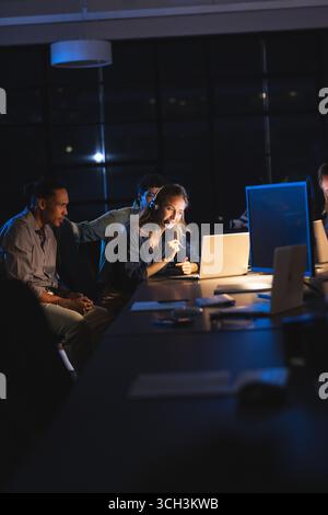 Diverse team collaborating at night in office, focused on laptop screens Stock Photo