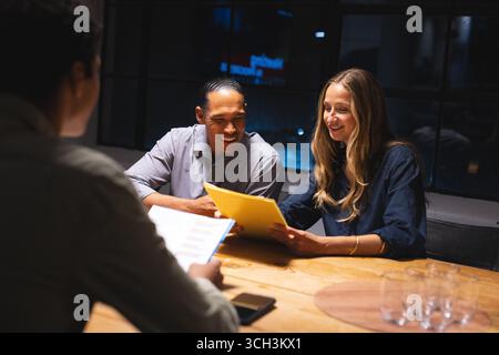 Discussing documents, diverse colleagues collaborating and smiling in night office meeting Stock Photo