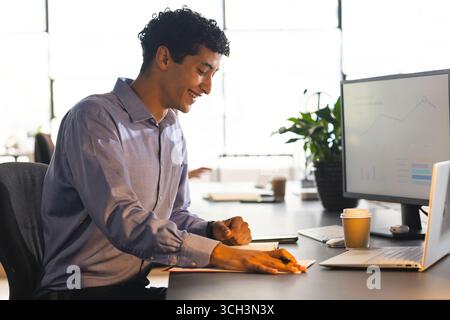 Smiling man at office desk analyzing data on computer, holding pen Stock Photo