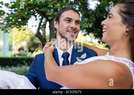 Groom in blue suit joyfully carrying bride outdoors, both smiling and celebrating Stock Photo