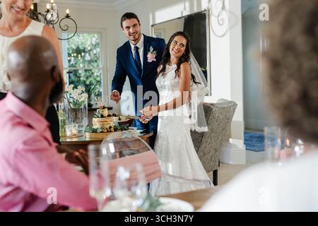 Bride and groom cutting cake, smiling at guests during wedding celebration indoors Stock Photo