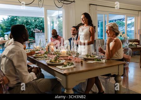Bride in white dress joyfully speaking to guests at elegant dinner table indoors Stock Photo
