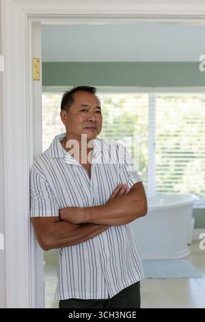 Senior Asian man standing with arms crossed, looking thoughtful in bright home Stock Photo