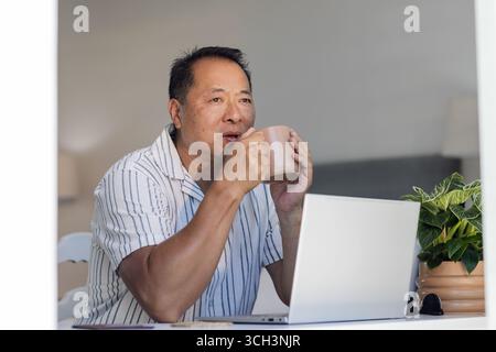 Senior Asian man enjoying coffee while using laptop at home, looking thoughtful Stock Photo