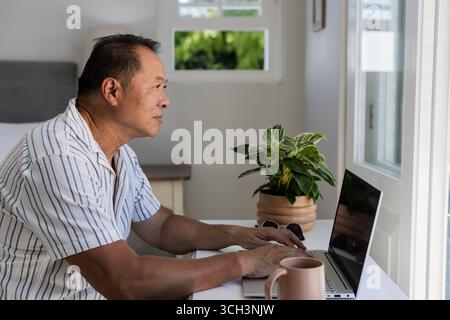 Senior man using laptop at home, focusing on work near window Stock Photo