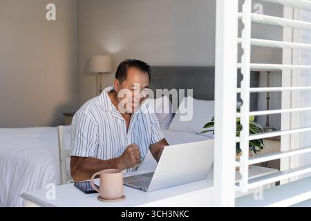 Senior Asian man celebrating success while video chatting on laptop at home office Stock Photo