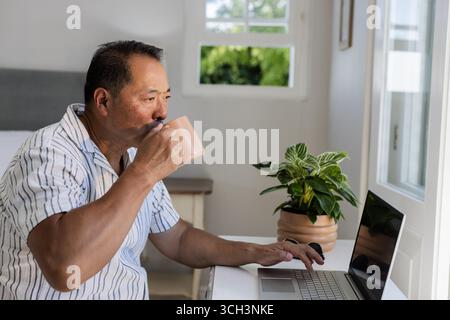 Senior Asian man drinking coffee while using laptop at home, enjoying morning routine Stock Photo