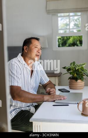 Senior Asian man sitting at desk with laptop, looking thoughtful at home Stock Photo