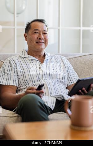Senior man relaxing on sofa, holding remote and tablet, enjoying leisure time Stock Photo
