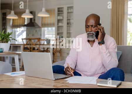 Senior man working from home, using laptop and talking on smartphone Stock Photo