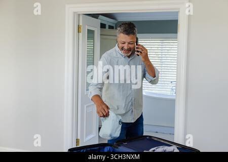 Senior man packing suitcase while talking on phone at home, looking cheerful Stock Photo