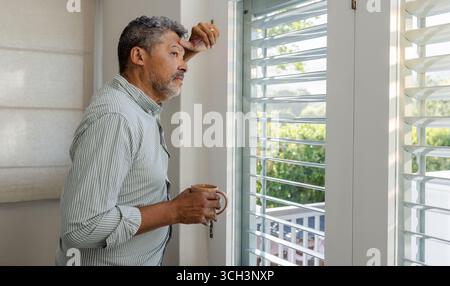 Senior man holding coffee cup, gazing thoughtfully out window at home Stock Photo