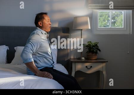 Asian man sitting on bed in bedroom, relaxing and enjoying morning sunlight Stock Photo