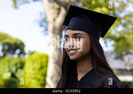Diverse woman standing and smiling in sunny park, with graduation cap and gown, copy space Stock Photo