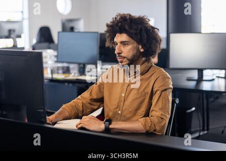 Young adult Asian man typing code at desk in open?plan office, with computer monitor and smartwatch Stock Photo