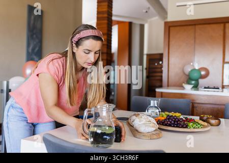 Woman photographing cutting board with bread and grapes using smartphone in kitchen, copy space Stock Photo