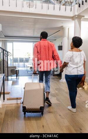 African American Couple entering modern home, carrying suitcase, holding hands, feeling excited Stock Photo