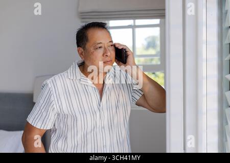 Senior man talking on smartphone while looking out window at home Stock Photo