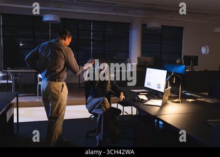 Colleagues discussing project in office at night, analyzing data on computer screen Stock Photo