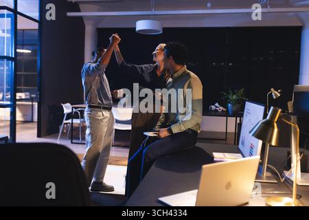 Celebrating success, diverse colleagues high-fiving in modern office at night Stock Photo