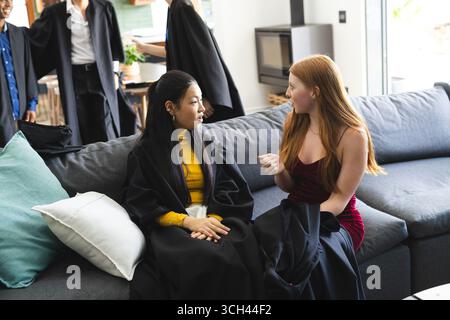 Teen friends in graduation gowns sitting on couch, talking and celebrating together Stock Photo